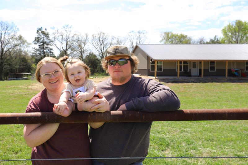 Husband, wife, and young child stand in front of their new home.