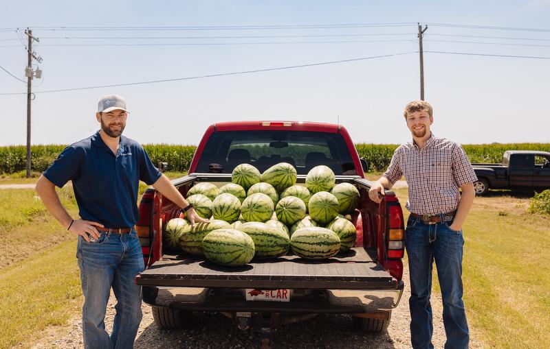 Two men stand behind red pickup truck full of watermelons.