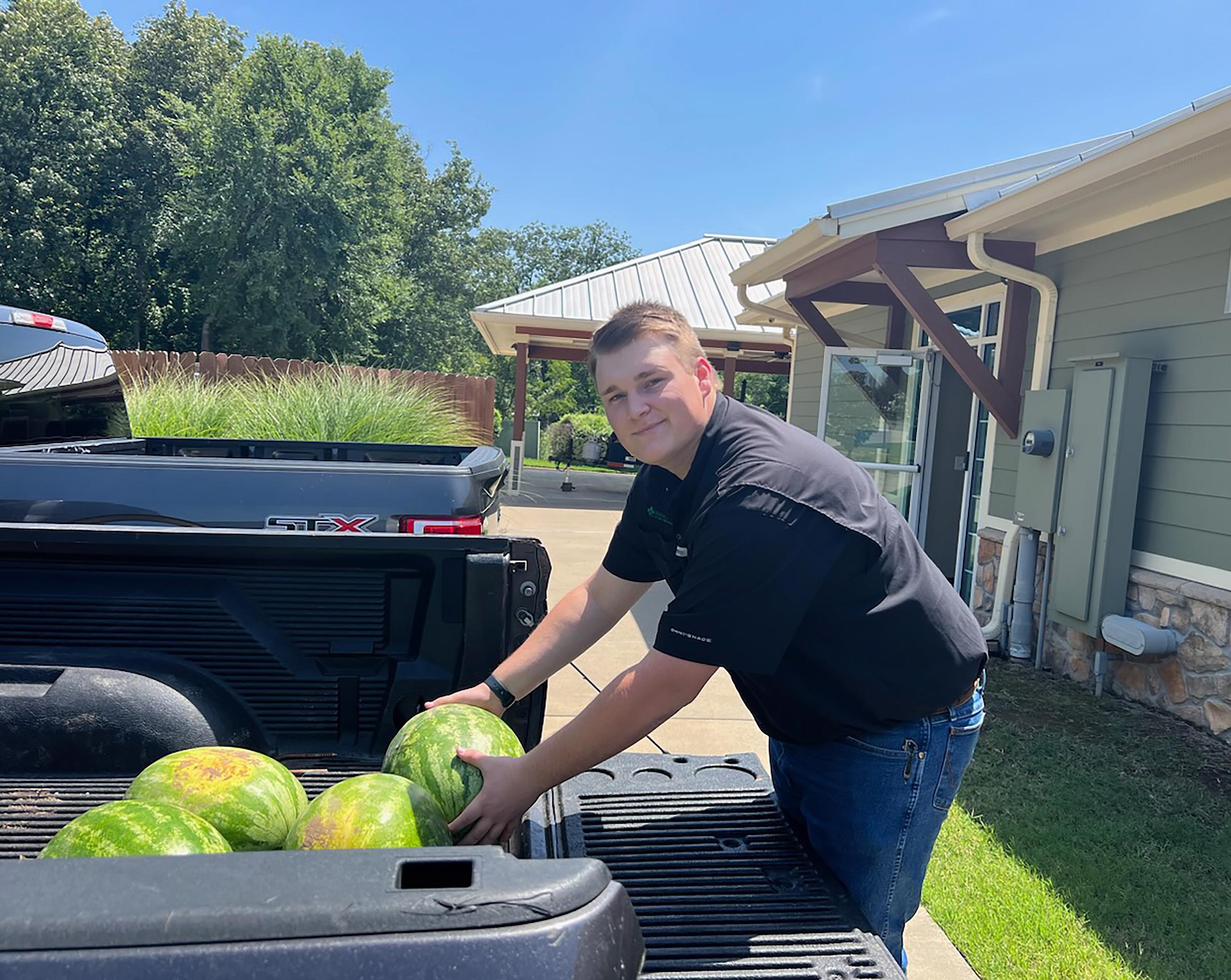 Austin Bell, Field Services Intern, loads watermelons into back of truck while smiling at camera